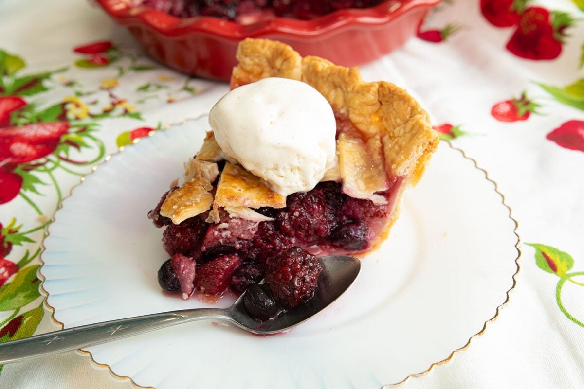 A slice of mixed berry pie, inspired by classic strawberry recipes, topped with vanilla ice cream sits on a white plate with a silver spoon. A pie dish and colorful tablecloth with fruit designs are in the background.