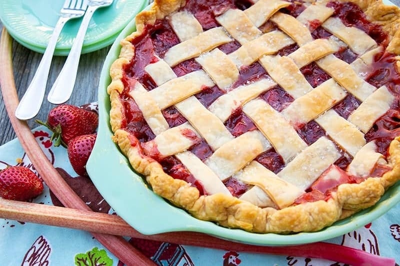A freshly baked strawberry rhubarb pie with a golden lattice crust sits on a table, surrounded by fresh strawberries, rhubarb stalks, and utensils on a colorful napkin—the perfect centerpiece for anyone seeking delicious Strawberry Recipes.