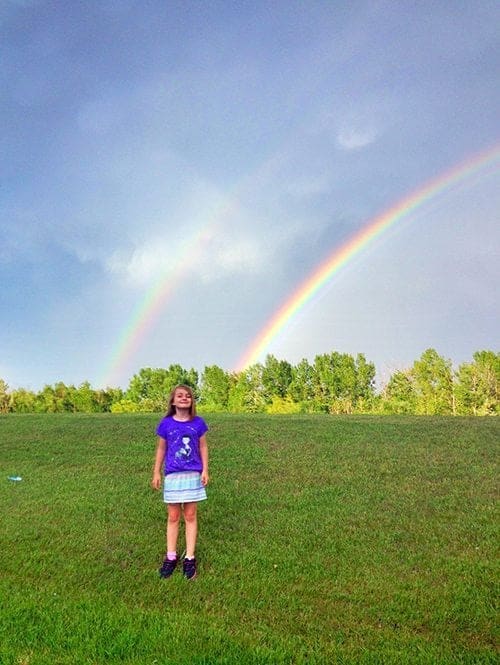 young girl in blue shirt and skirt standing in the field with rainbow on background