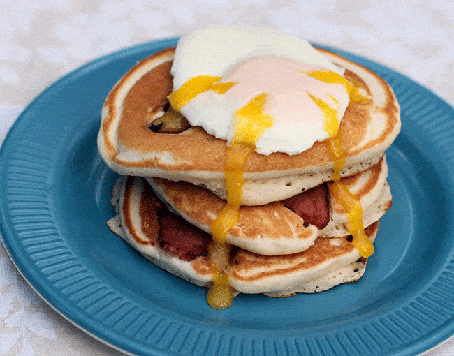 three pieces bacon pancakes with fried egg on top, letting the luscious yolk drip down all over the bacon pancakes
