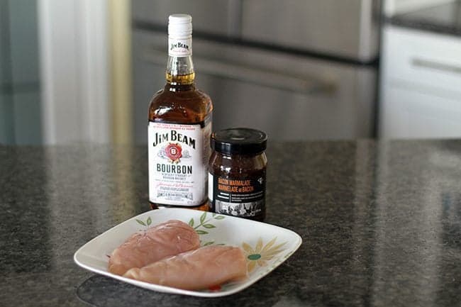 A bottle of bourbon, jar of bacon jam and two pieces of chicken breast in a plate