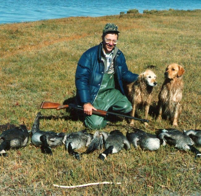 fallen ducks in front of man holding his rifle and his two dogs