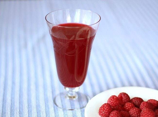 a glass of Raspberry Cordial and a white plate with fresh raspberries on a blue stripe background