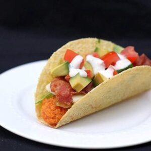 Chicken Tacos on a white plate loaded with diced avocados, tomatoes, shredded lettuce and bacon