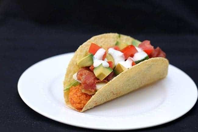 Chicken Tacos on a white plate loaded with diced avocados, tomatoes, shredded lettuce and bacon