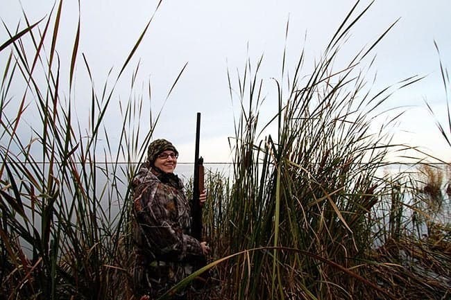 woman wearing duck hunting gear, standing and holding her riffle 