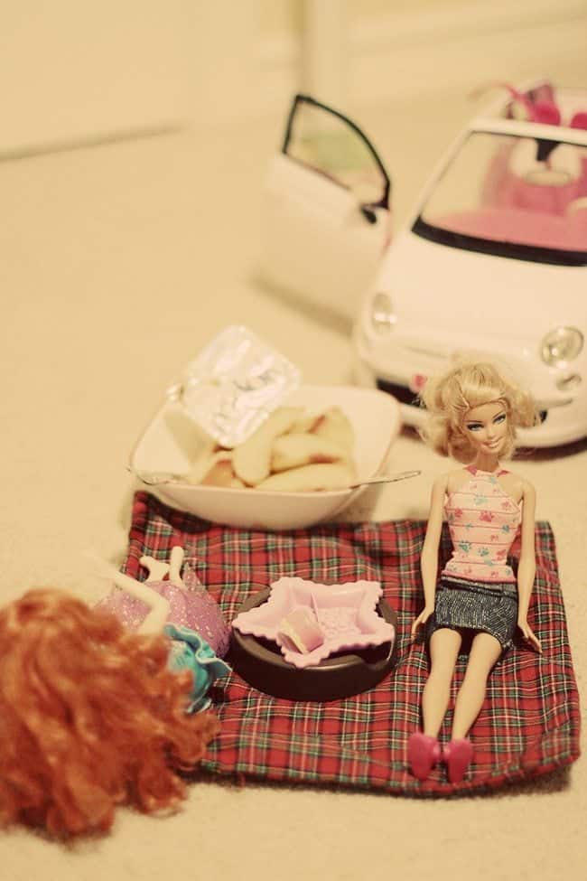 close up of barbie dolls sitting and a plate bowl with Yopa and slices of apple beside it