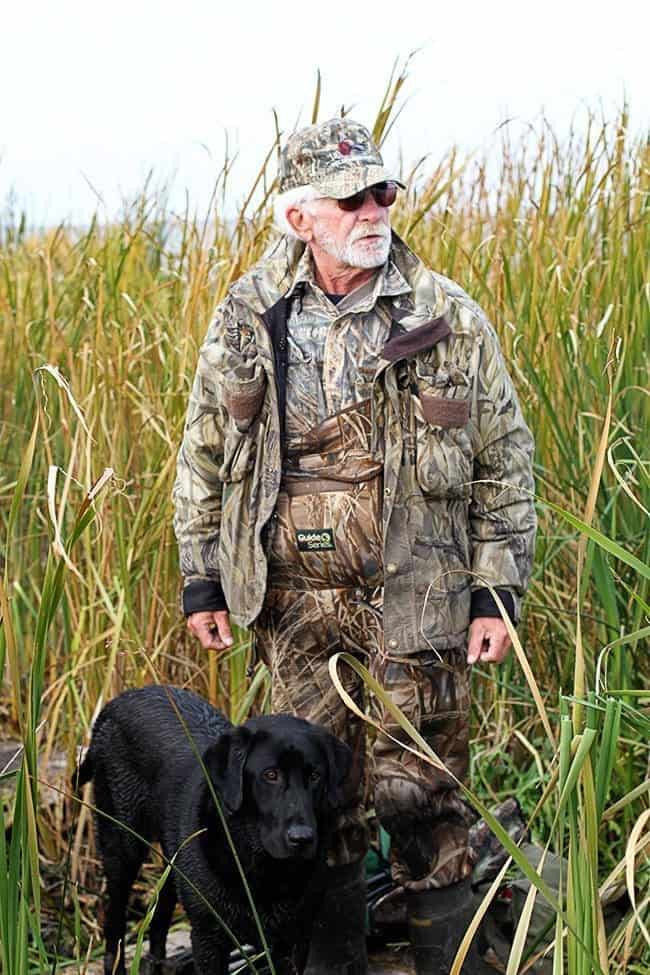 man wearing duck hunting gear standing in tall grasses with the big black dog