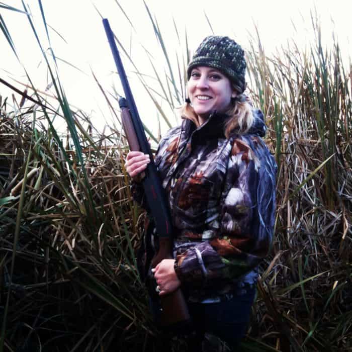 woman wearing her duck hunting gear, holding a riffle while standing within tall grasses