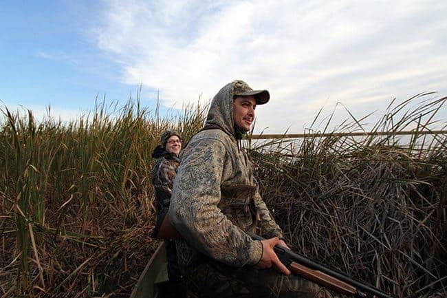 man holding riffle and woman both wearing duck hunting gear in tall grasses
