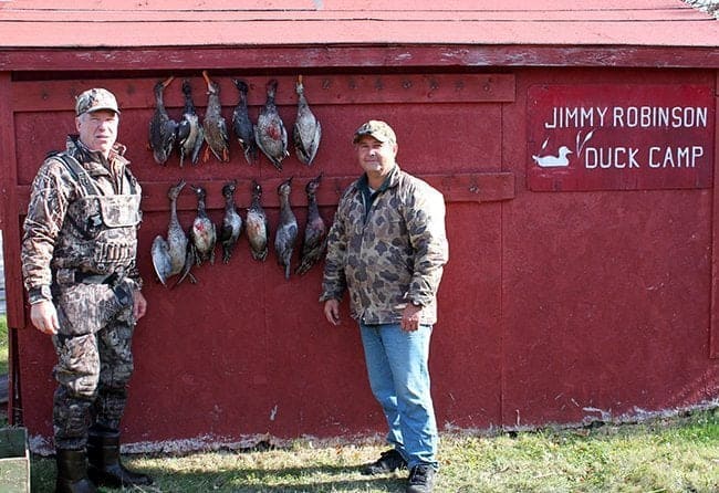two men wearing duck hunting gears with hanging ducks in their background