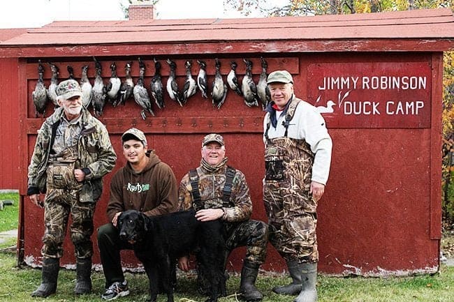 four men and a black dog with hanging ducks background