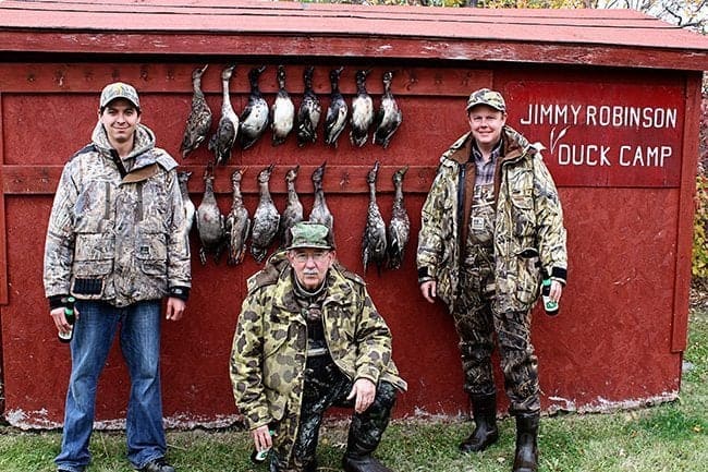 three men wearing duck hunting gears with hanging ducks background