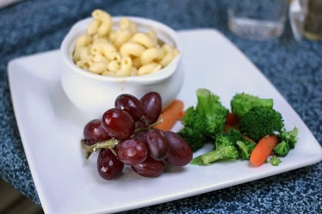 kids meals in a white plate with macaroni in a small bowl, fruits and vegetables on the side