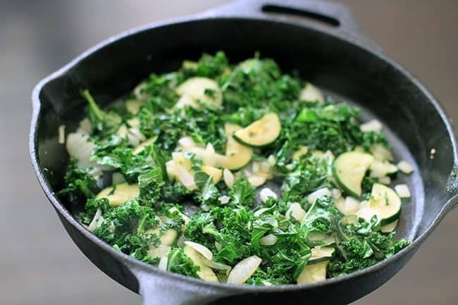 Large black pan with onions, kale, vegetables, and zucchini sliced in it.