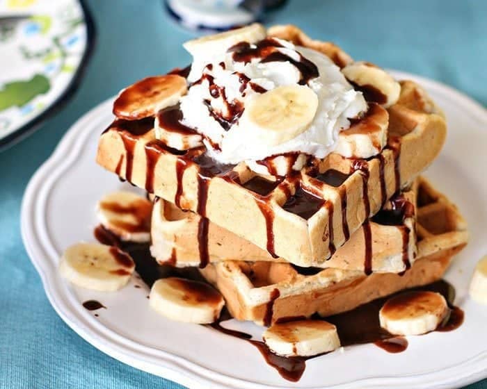 Close up Stack of Banana Waffles in a White Plate, topped with whipped cream, fresh bananas and Nutella sauce