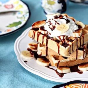 Close up Stack of Banana Waffles in a White Plate, topped with whipped cream, fresh bananas and Nutella sauce
