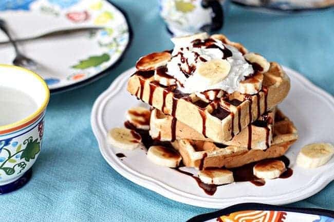 Stack of Banana Waffles in a White Plate, topped with whipped cream, fresh bananas and Nutella sauce