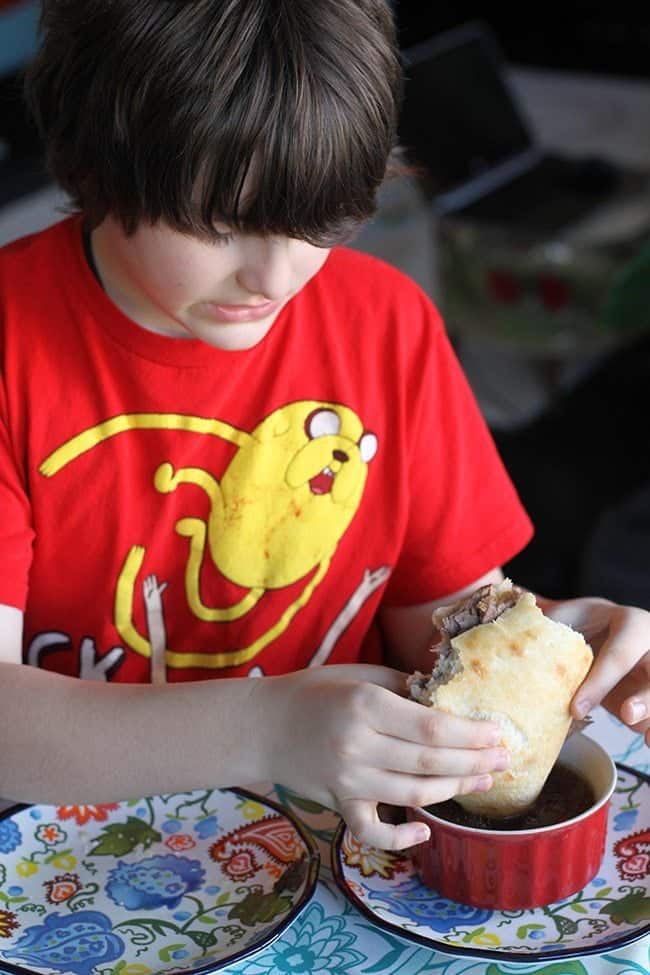 boy wearing red shirt holding sandwich and dipping it to crock pot beef dip