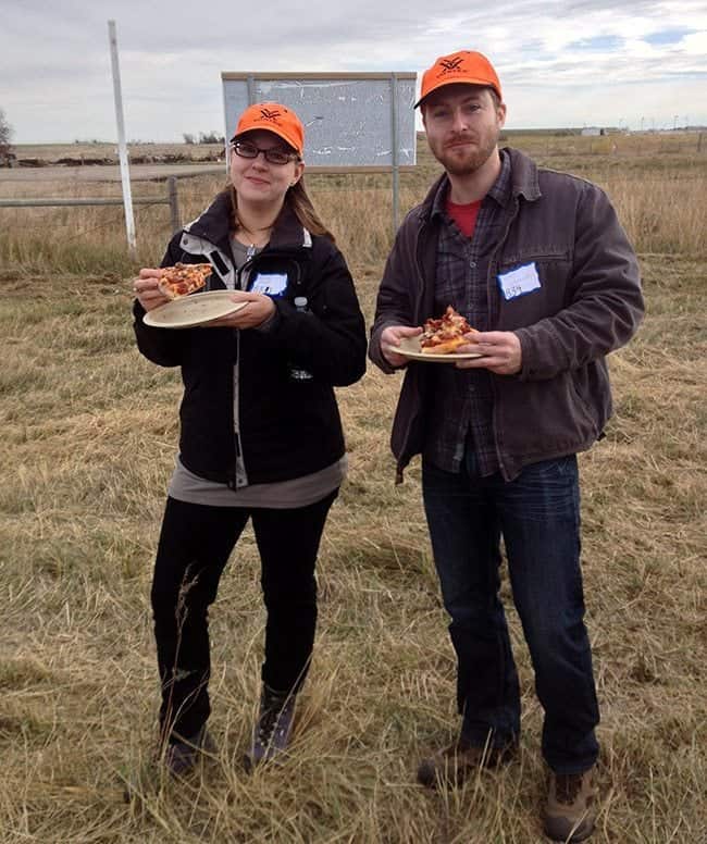 man and woman wear matching hats, standing and holding plate with a slice of pizza