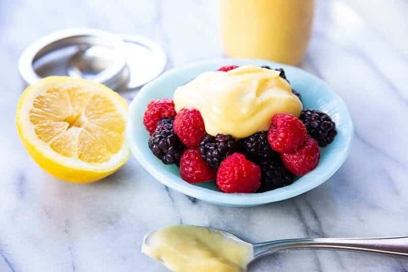 Lemon curd on top of raspberries and blackberries in a small Pyrex bowl, a slice of lemon and spoon with curd beside it