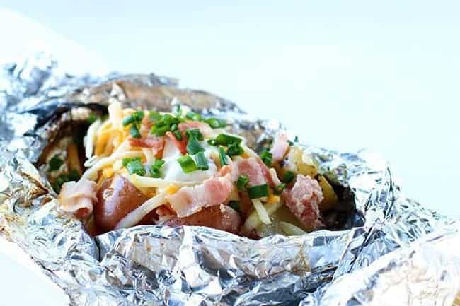 close up Campfire Grilled Loaded Baked Potatoes in a tinfoil topped with onion, cheese, sour cream, salt and pepper