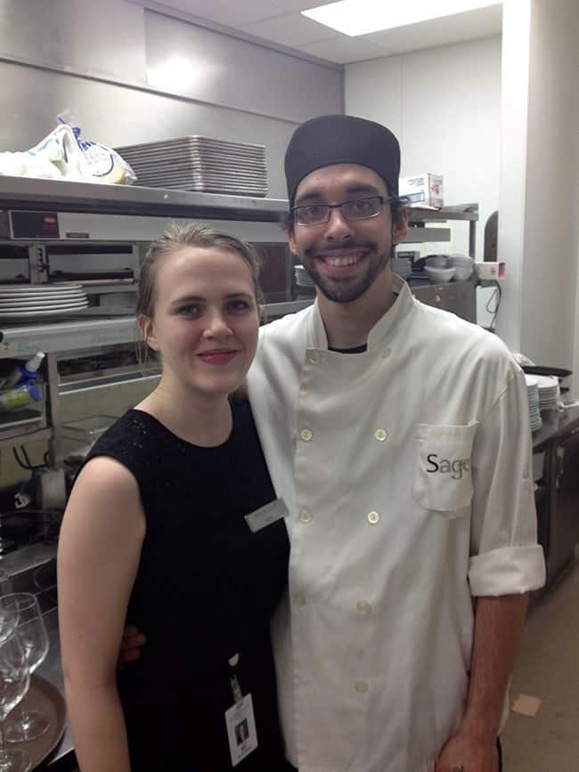 a lady black dress and a chef with beard at the kitchen