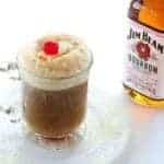 close up of glass of Bourbon Root Beer Ice Cream Floats with whipped cream and cherry. A bottle of Jim Beam bourbon at the back.