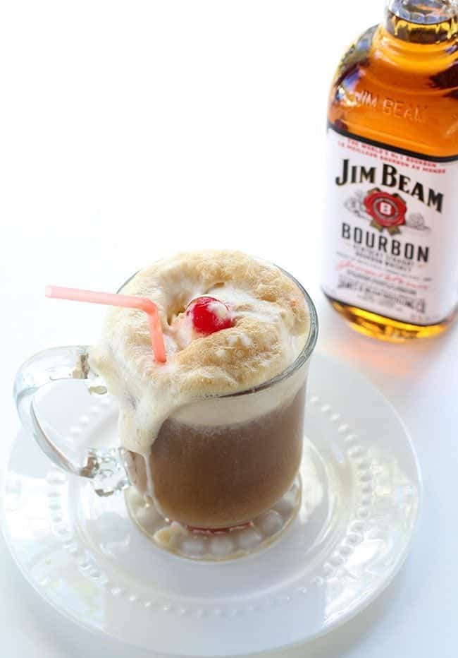 close up of glass of Bourbon Root Beer Ice Cream Floats with whipped cream and cherry. A bottle of Jim Beam bourbon at the back.