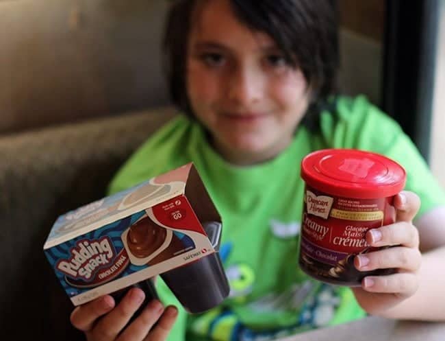 boy wearing a green shirt, holding cups of chocolate pudding snack and can of chocolate icing