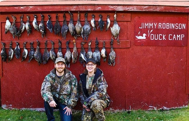 man and woman in their hunting gear with hunted ducks hanging on their background