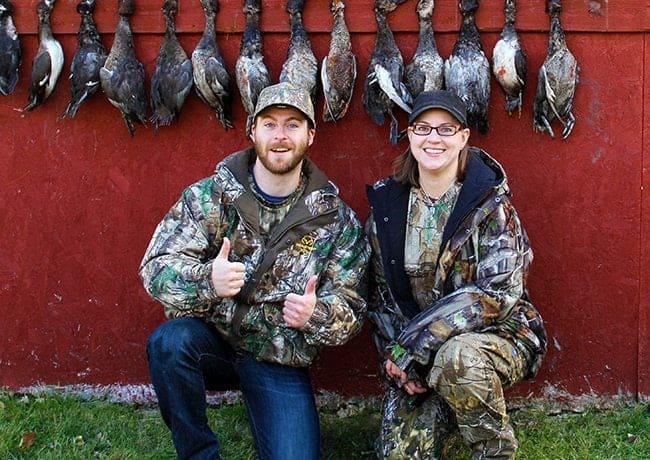 man in thumbs up and woman in their hunting gear with hunted ducks hanging on their background