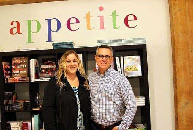 lady in black coat and man in polo in front of cookbooks shelves