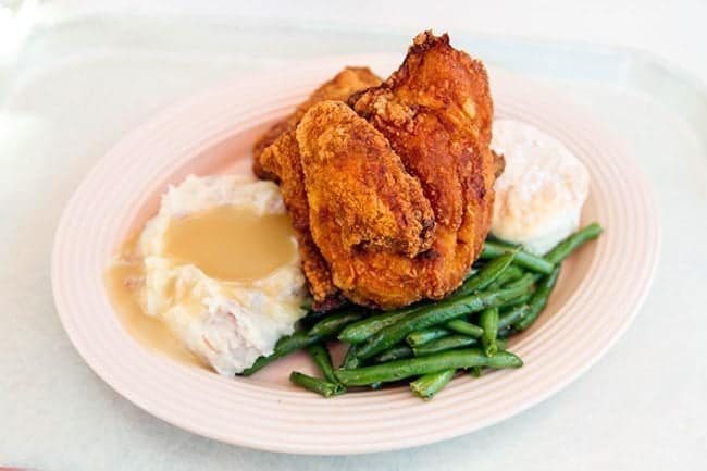 white plate with 3 pieces of chicken, a biscuit, a pile of potatoes and green beans