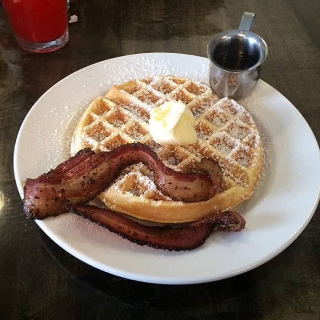 Peppered bacon and a waffle with butter on top, a cup of pancake syrup in a white plate