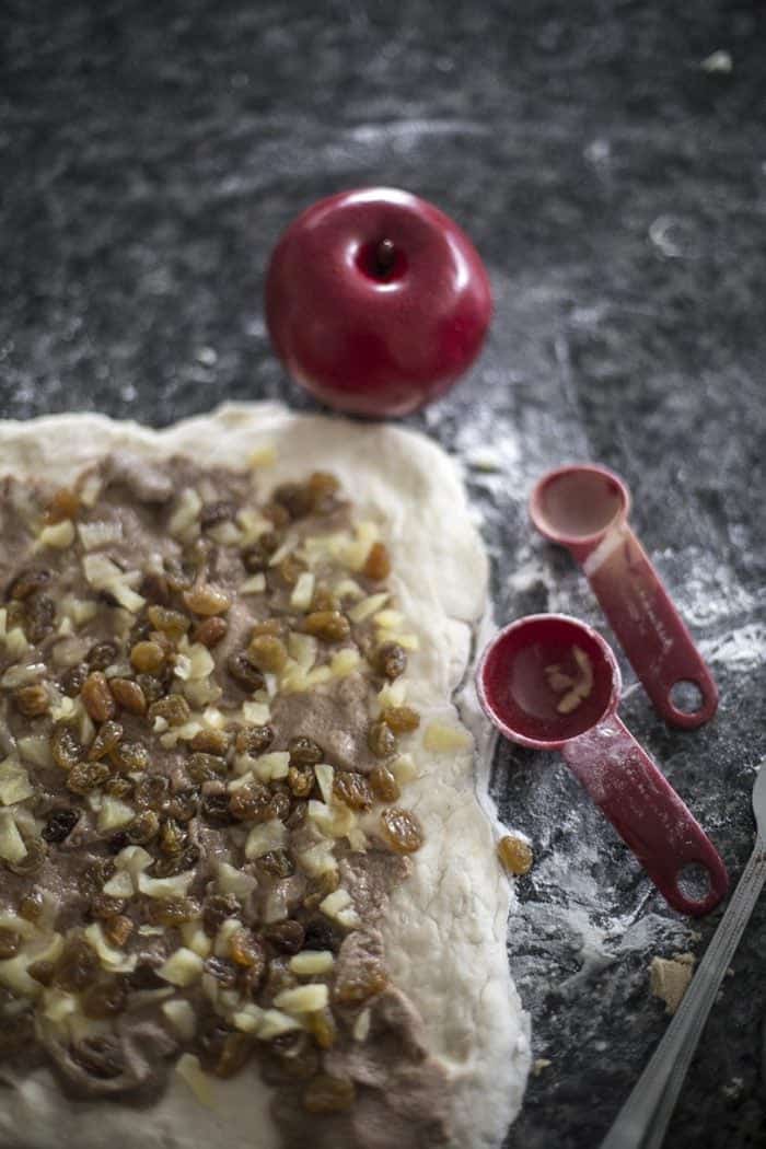 Apple Cinnamon Buns dough rolled into rectangular shape added with filling, red apple and measuring spoons on side