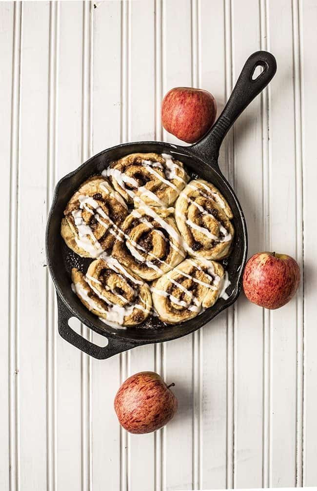 top down shot of 6 pieces Glazed Apple Pie Cinnamon Buns in Skillet with 3 fresh apples on white background