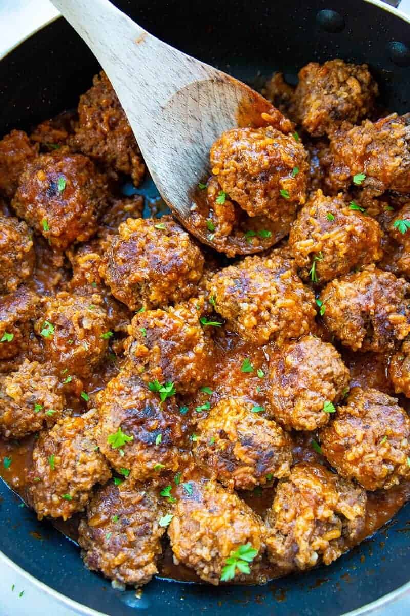 close up porcupine meatballs in a large black skillet with a wooden spoon and some teared parsley leaves