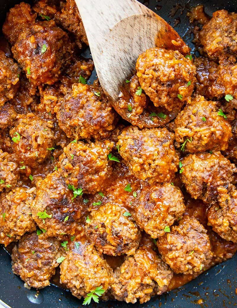 close up porcupine meatballs in a large black skillet with a wooden spoon and some teared parsley leaves