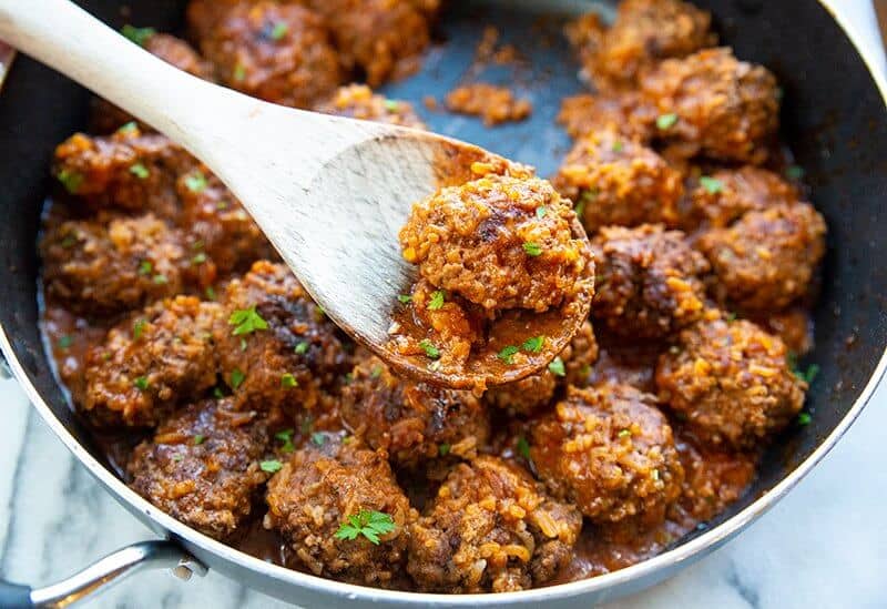 porcupine meatballs in a large black skillet with a wooden spoon and some teared parsley leaves