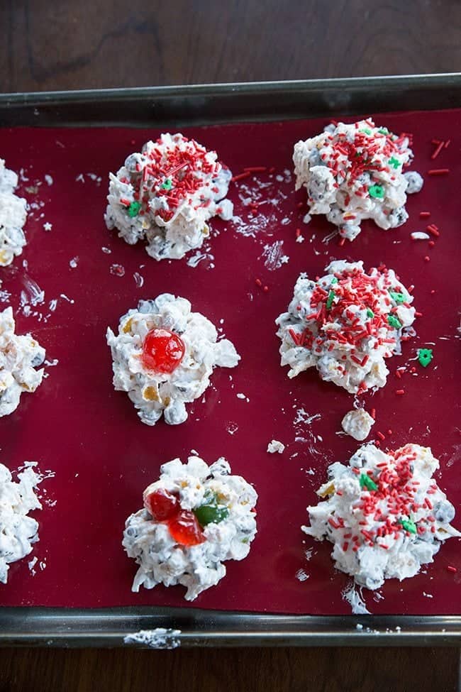 meringues with corn flakes, chocolate chips, coconut and cherries on top in a parchment lined baked sheet