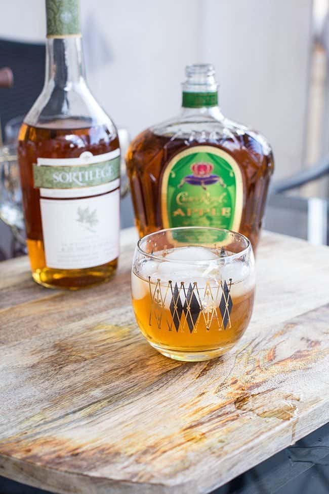 a glass of Log Cabin Cocktail with a bottle of Sortilege and apple cider on background