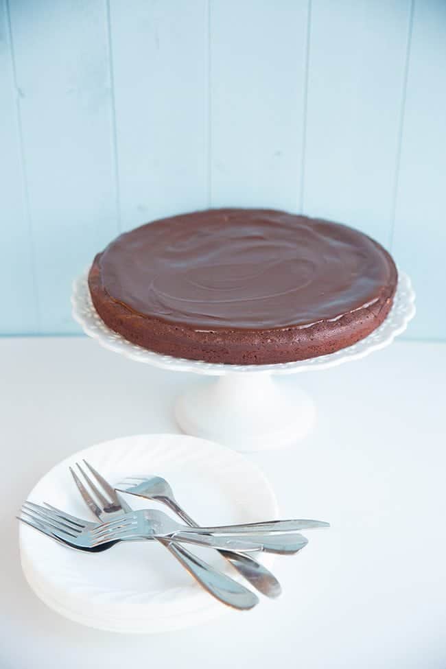 Flourless Chocolate Cake in a white cake stand, dessert plates and forks beside it