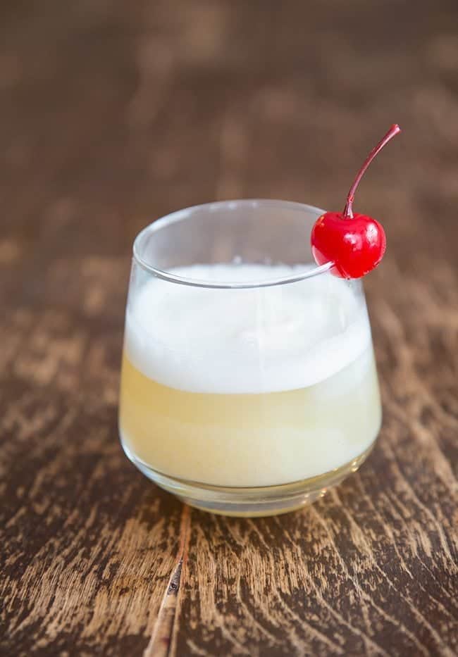 A glass of Smoky Whisky Sour with egg white on top and Garnish with Cherry on Wood background