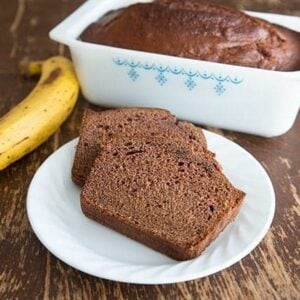  Slices of Chocolate Banana Bread in White plate and a loaf in baking pan, ripe yellow banana on side