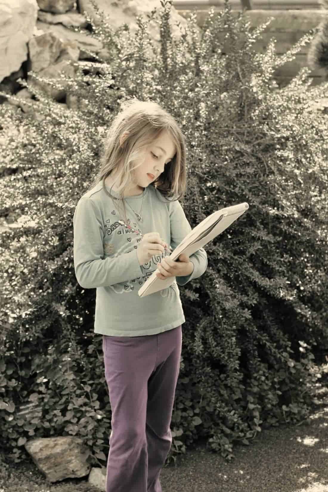 little girl standing, holding her sketchpad and pencil in the garden