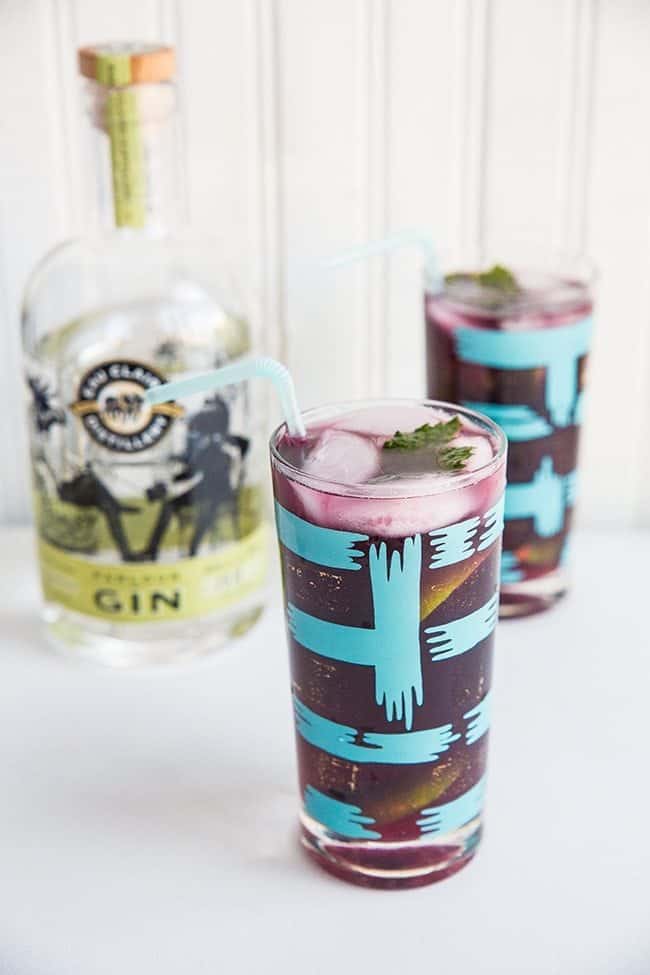 Tall turquoise cocktail glass with Blueberry Mint Gin Fizz and a bottle of Eau Claire Distillery on the background