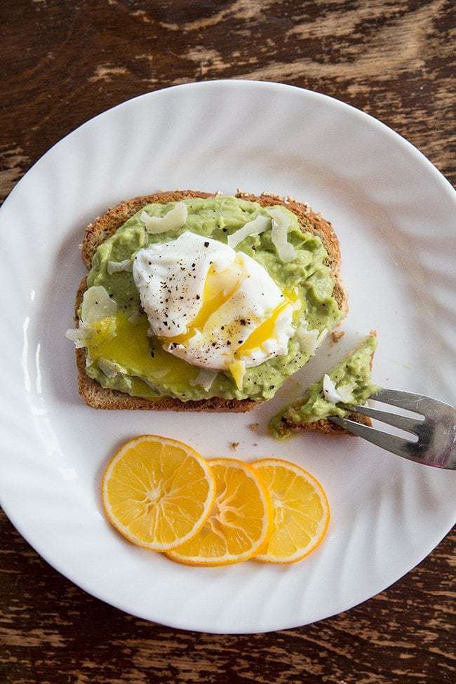 white plate with 3 lemon slices and toasted bread slice spread with mash avocado topped with poached egg and Parmesan cheese