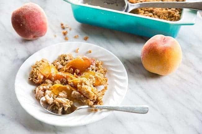 Peach Crisp in a white plate with spoon, peach fruit and blue baking dish with crisp on a marble background