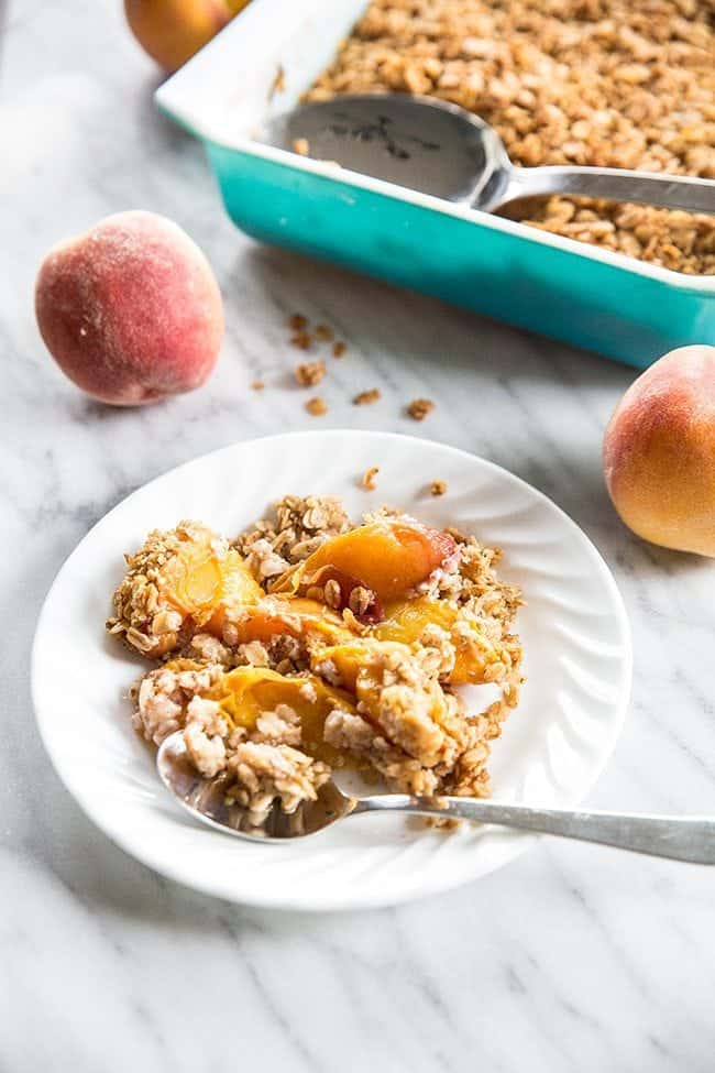 Peach Crisp in a white plate with spoon, blue pan with oat mixtures and peach fruit on a marble background